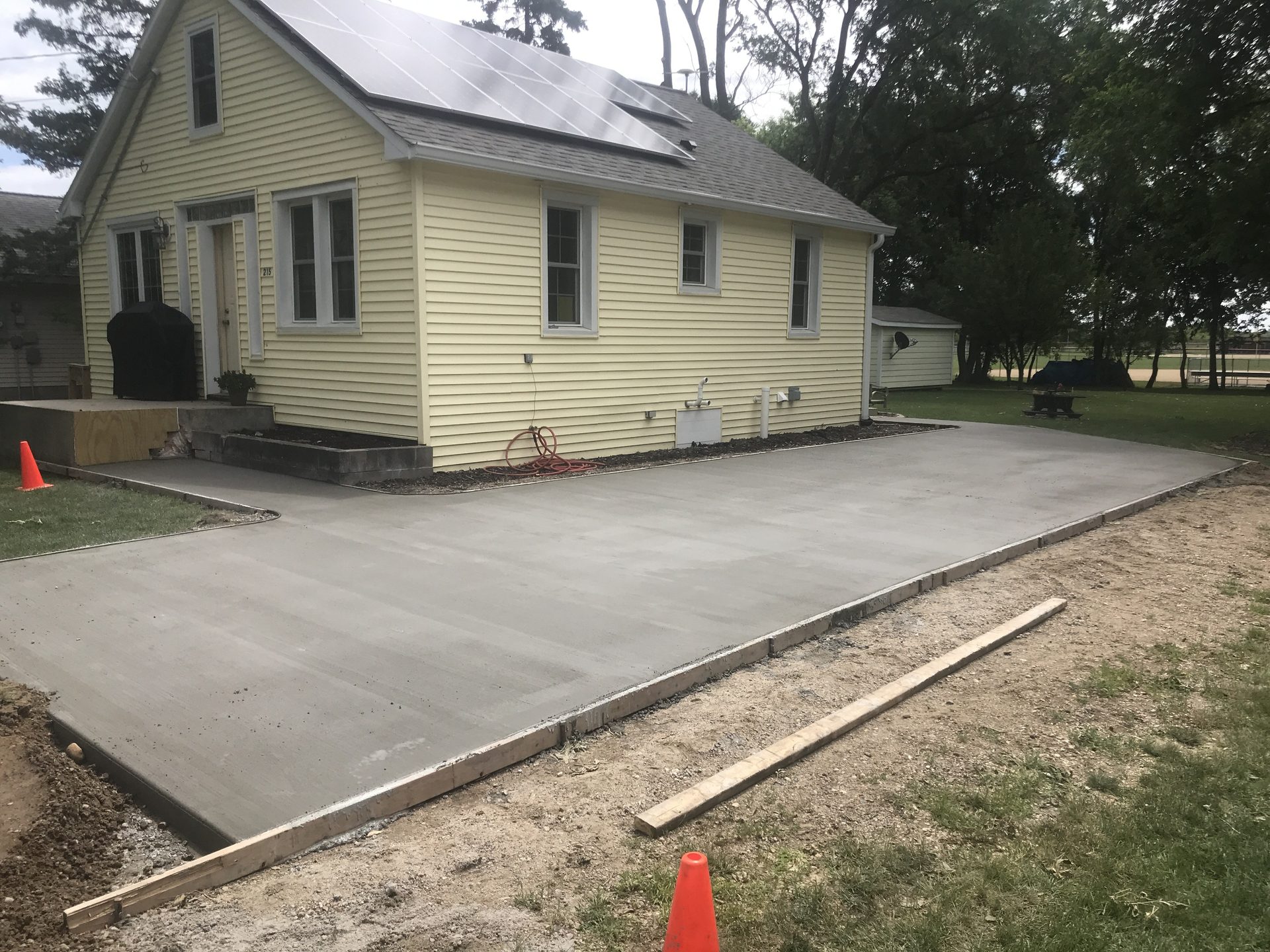 Newly poured concrete driveway alongside cream-colored ranch home with solar panels, showing finished concrete pad and surrounding site preparation.
