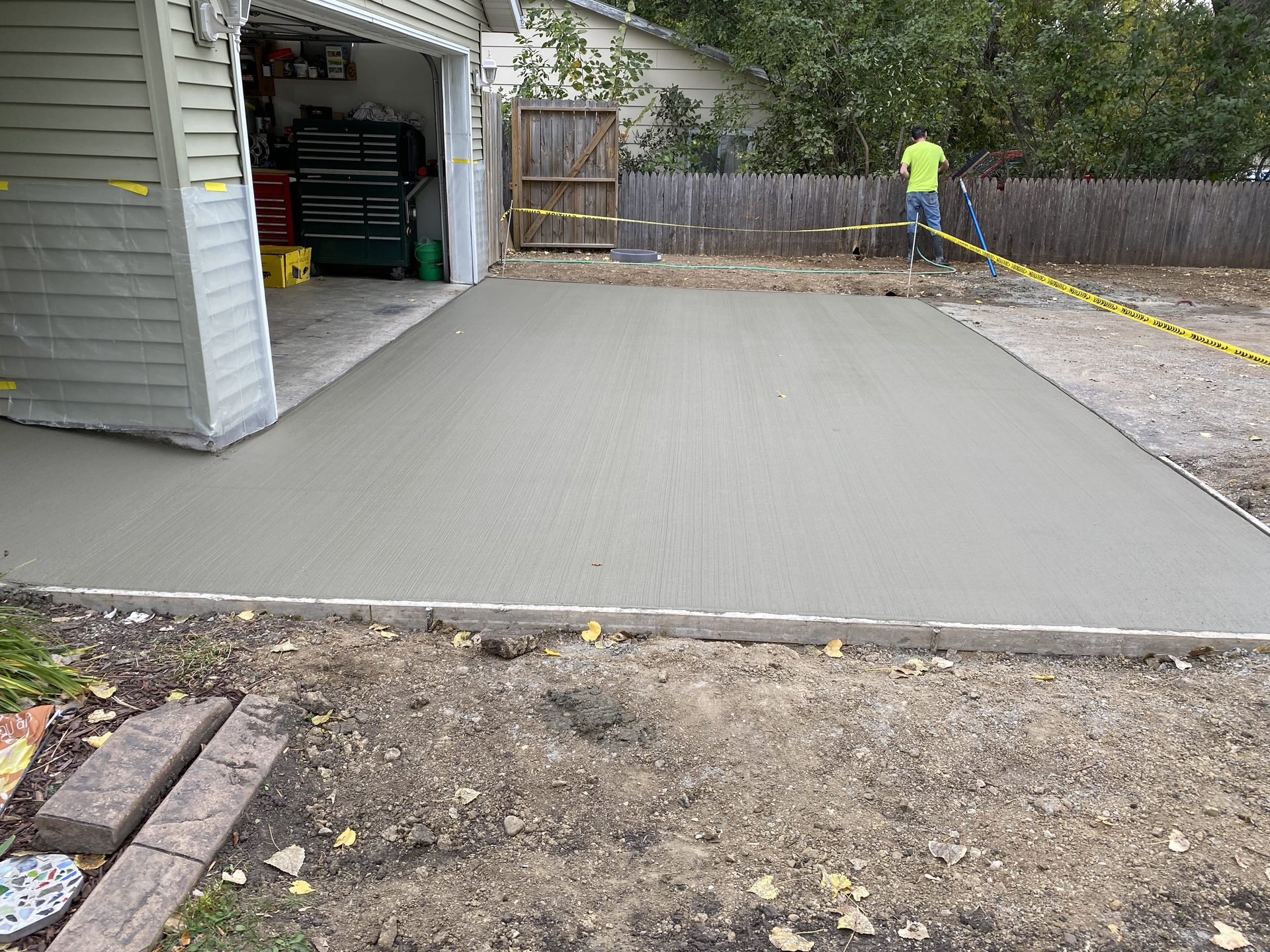 Freshly poured concrete driveway adjacent to garage, with worker and caution tape visible in background.