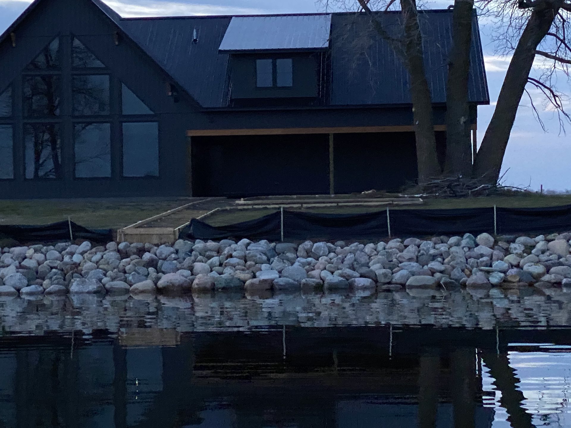 Modern dark-clad residential home with large windows overlooking a lake, featuring a stone riprap shoreline stabilization and wooden dock structure.