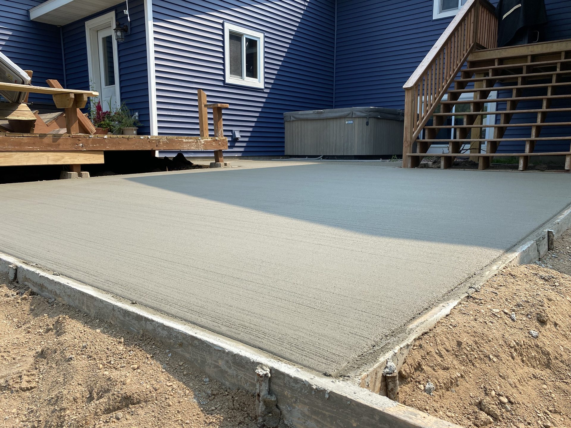 Newly finished concrete patio pad adjacent to blue-sided residential home with wooden deck and stairs visible.