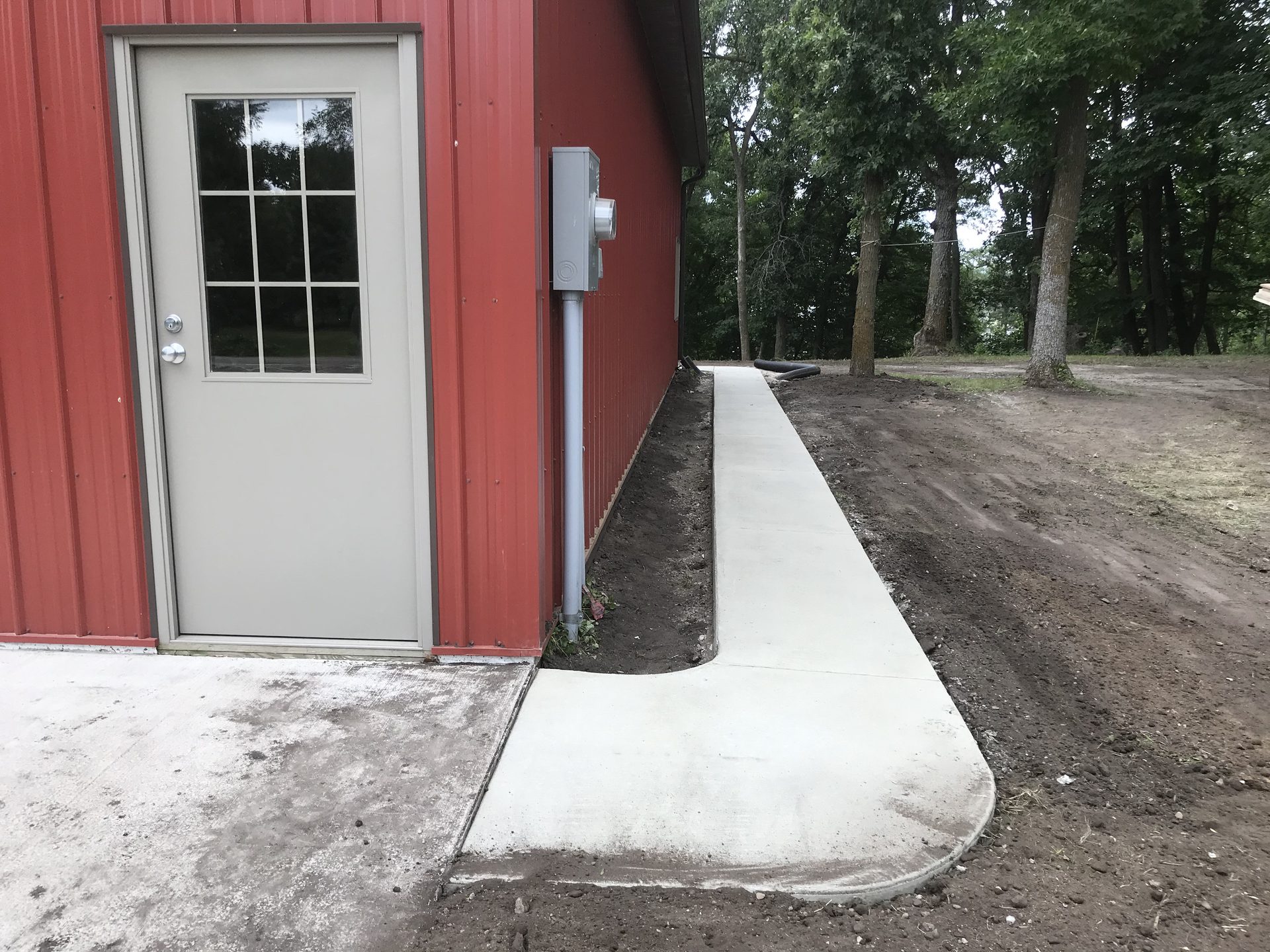 Newly installed concrete walkway alongside red metal building with white door and surrounding landscaping.