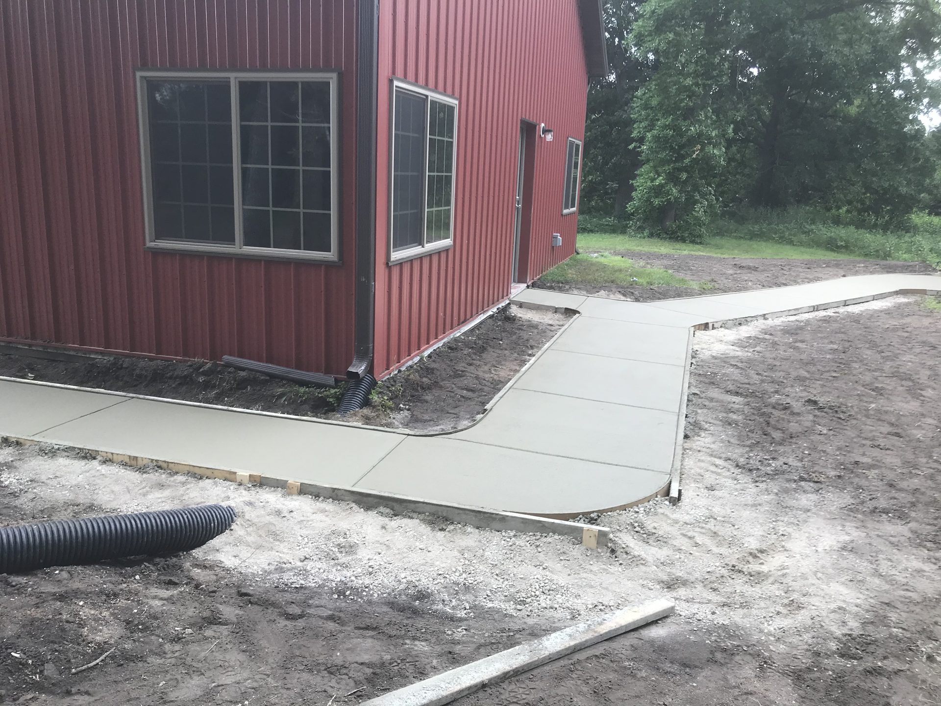 Concrete driveway and foundation pad installation alongside red metal building, with drainage pipe and gravel staging visible.