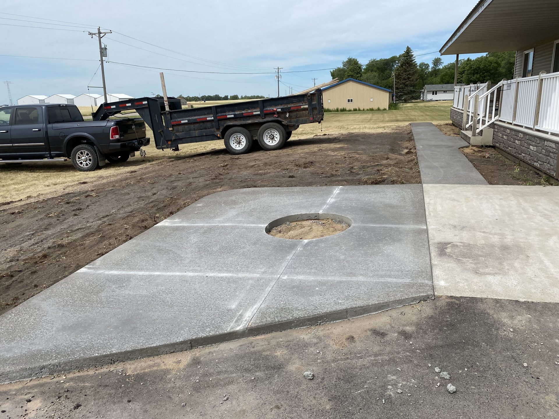 New concrete driveway with central drain opening, partially completed with dumptruck and equipment visible on rural jobsite.