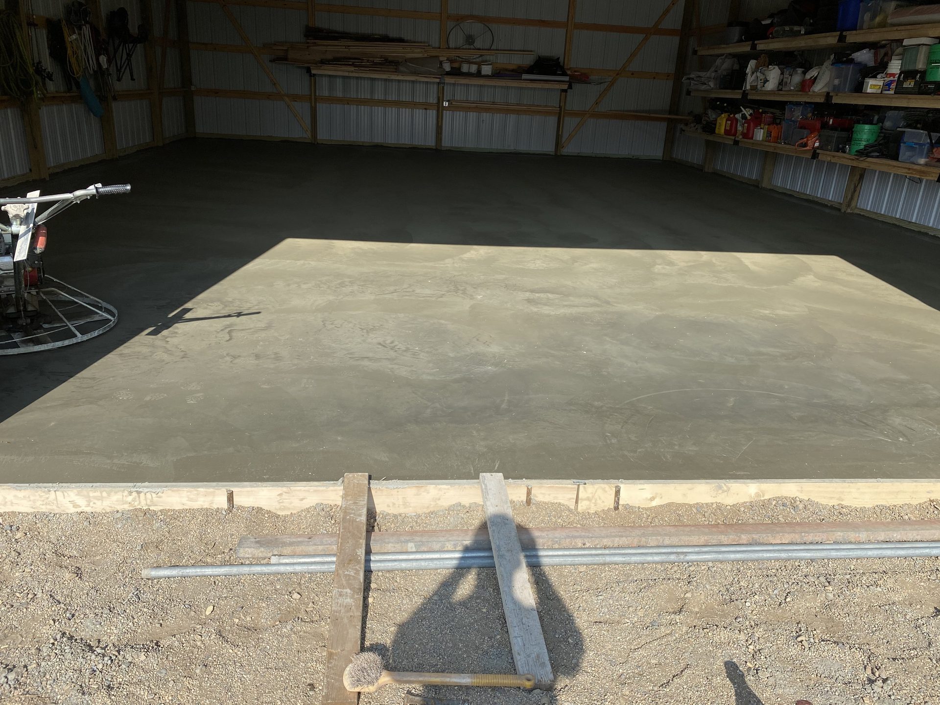 Fresh concrete slab in progress inside a metal-frame garage or warehouse, with finishing tools and jobsite equipment visible.