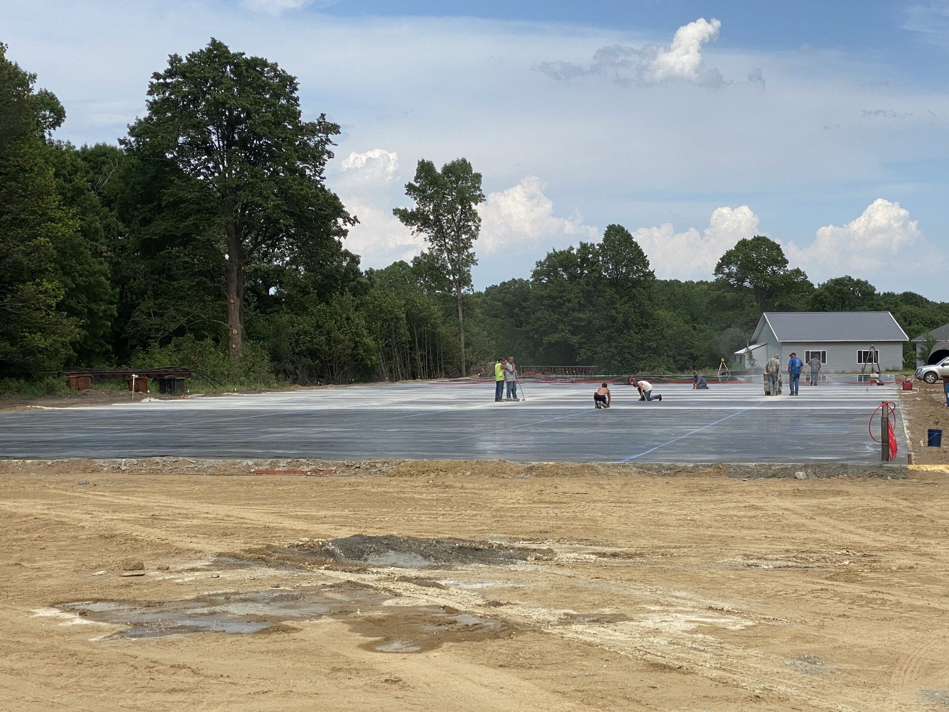 Concrete foundation slab for residential construction with workers finishing the surface, surrounded by cleared land and forest.