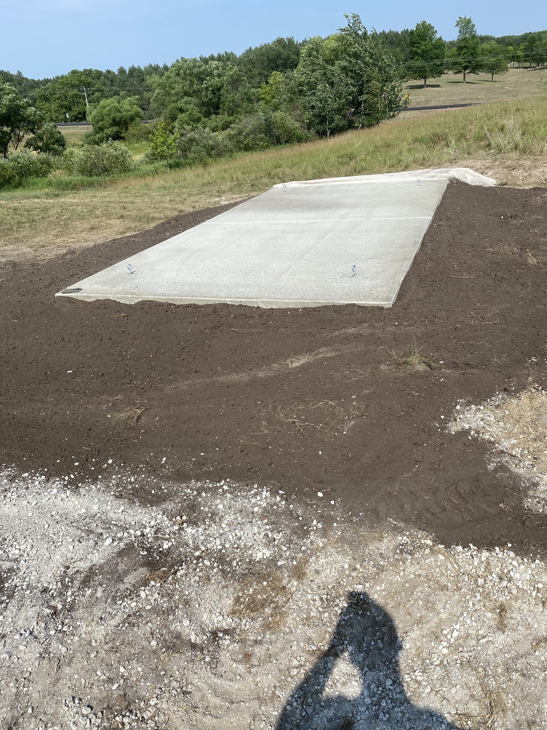 Newly poured concrete pad on rural jobsite with gravel base and surrounding earthwork, forested hills visible beyond.