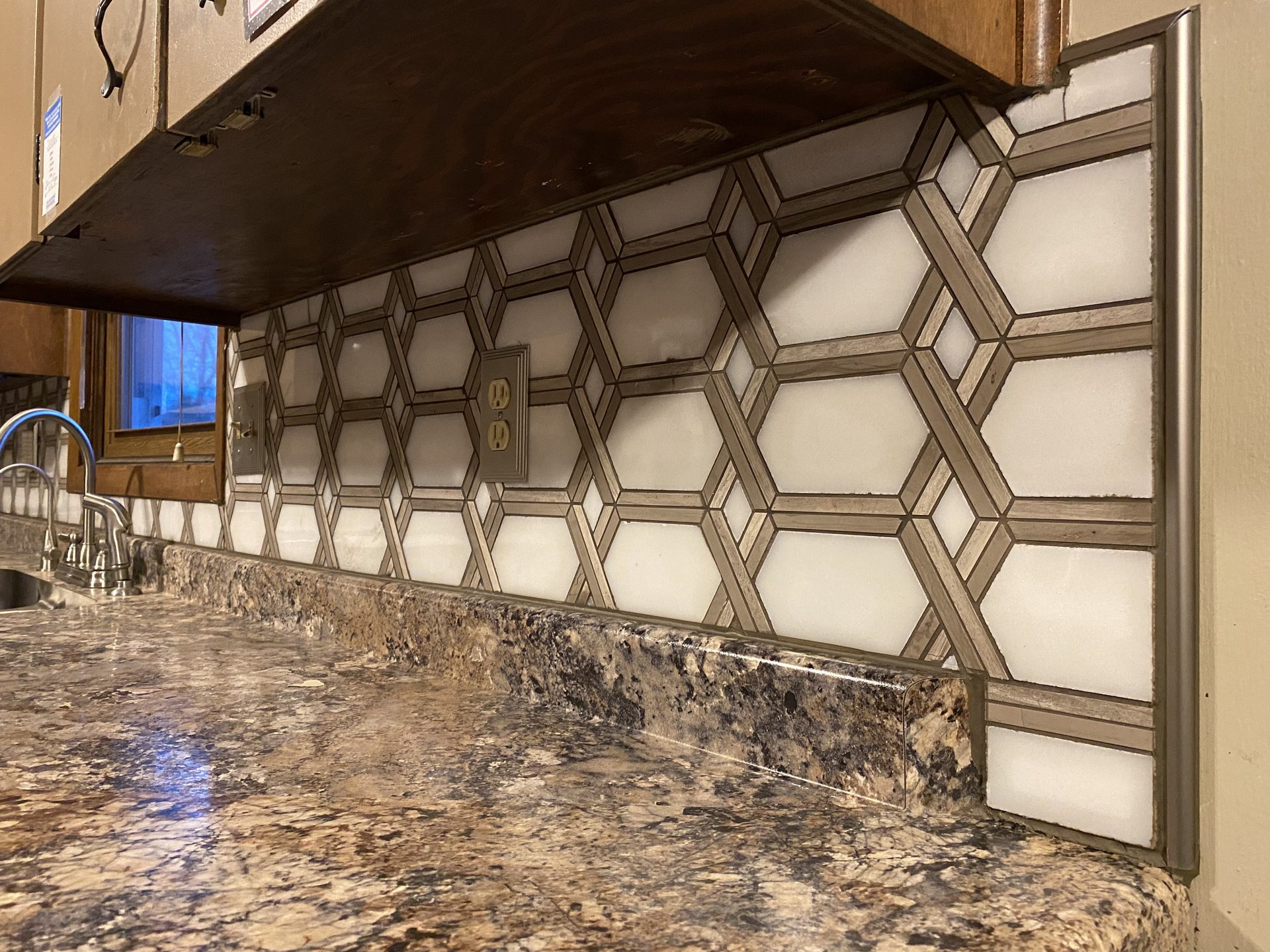 Kitchen backsplash installation featuring geometric patterned tile with bronze metallic grout lines above granite countertop.