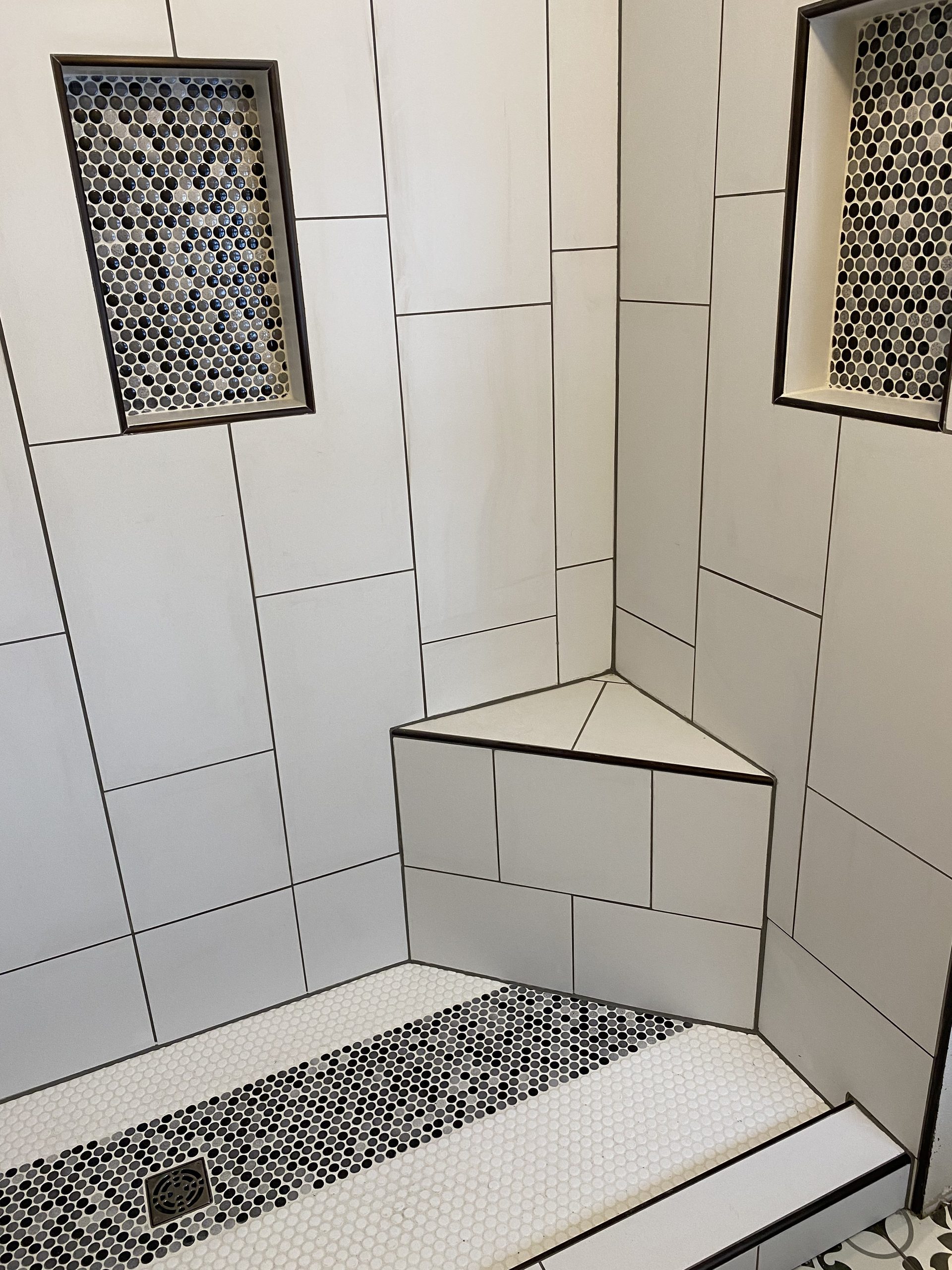 Completed tile shower with large format subway tiles, black grout lines, mosaic penny tile floor strip, corner bench, and framed niche details.