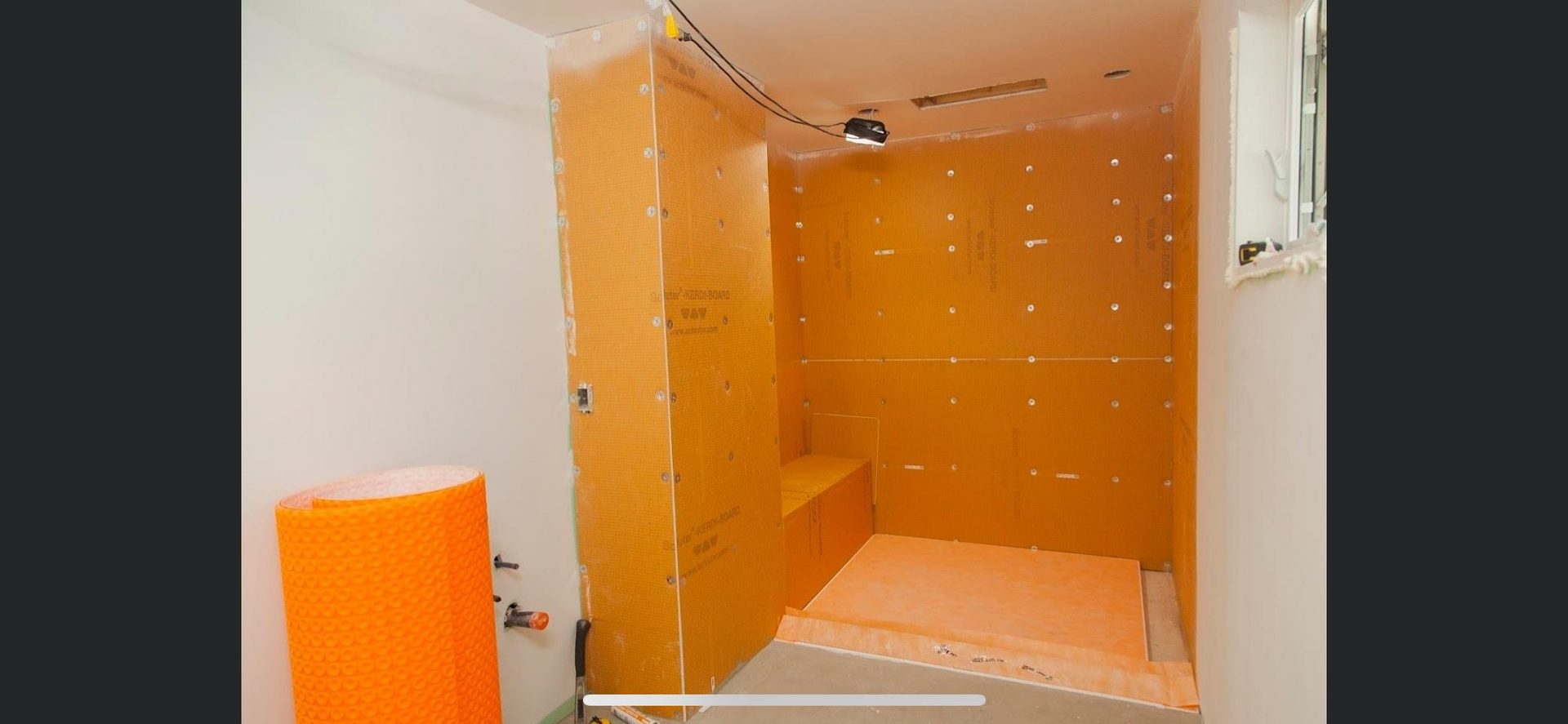Bathroom tile shower installation in progress, showing orange waterproofing membrane applied to framed walls with fixture rough-ins visible.