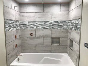 Completed tile shower surround with gray subway and mosaic accent tile, white bathtub visible.
